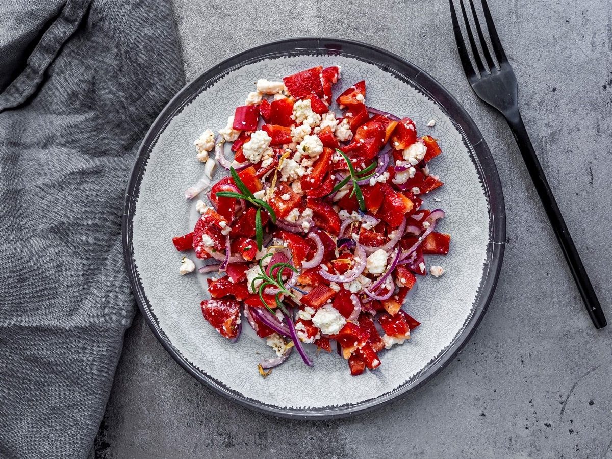 Ein weißer Teller auf grauem Untergrund mit Paprika-Salat mit Feta