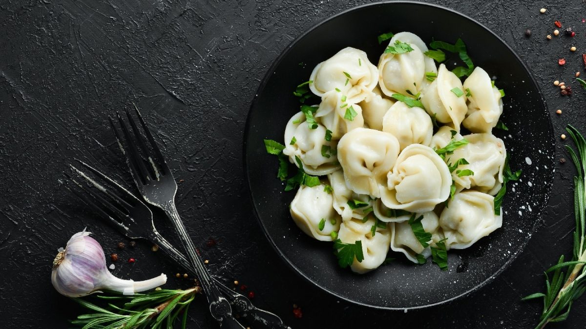 Ein schwarzer Teller mit Pelmeni auf einem dunklen Untergrund von oben fotografiert. Daneben Knoblauch, Pfeffer, Kräuter und schwarzes Besteck.