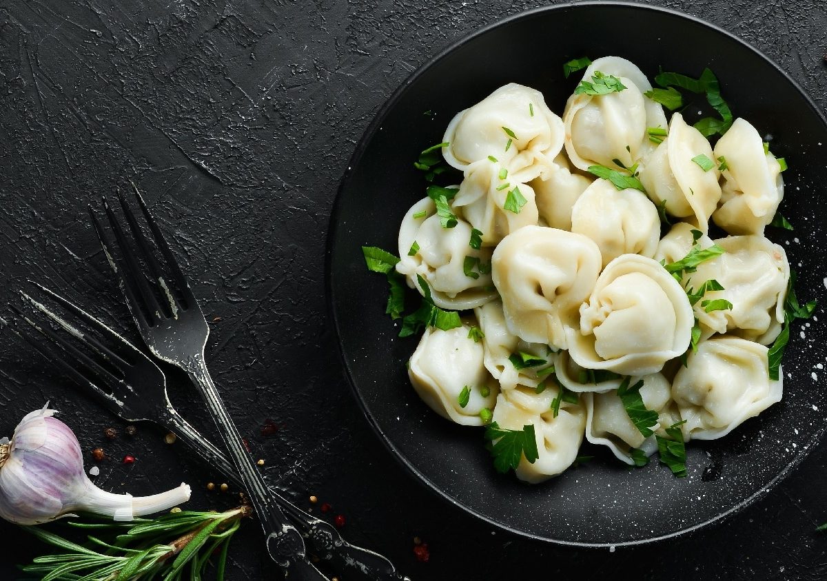 Ein schwarzer Teller mit Pelmeni auf einem dunklen Untergrund von oben fotografiert. Daneben Knoblauch, Pfeffer, Kräuter und schwarzes Besteck.