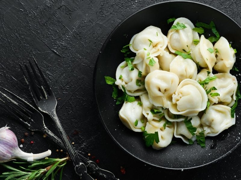 Ein schwarzer Teller mit Pelmeni auf einem dunklen Untergrund von oben fotografiert. Daneben Knoblauch, Pfeffer, Kräuter und schwarzes Besteck.