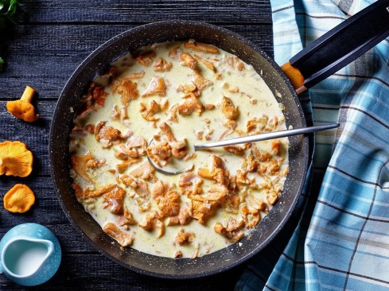 Eine Pfanne mit einer Pfifferling-Sauce steht auf einem Untergrund aus Holz. Drum herum liegen frische Pfifferlinge, Petersilie sowie ein Kännchen Sahne