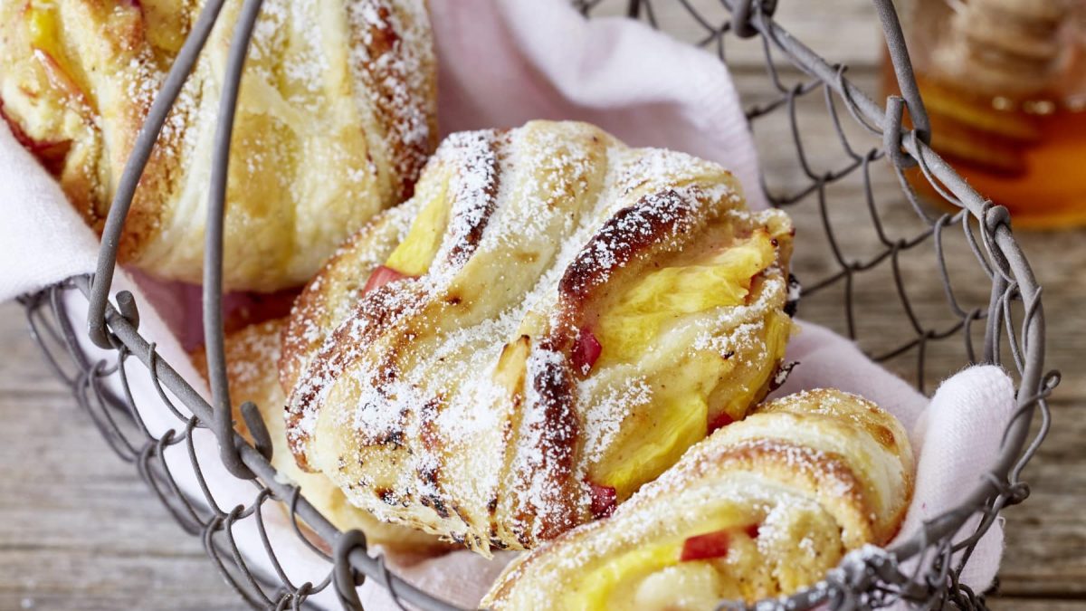 Drei Pfirsich-Schnecken mit Puderzucker auf heller Serviette in einem Metallkörbchen vor Holzhintergrund.