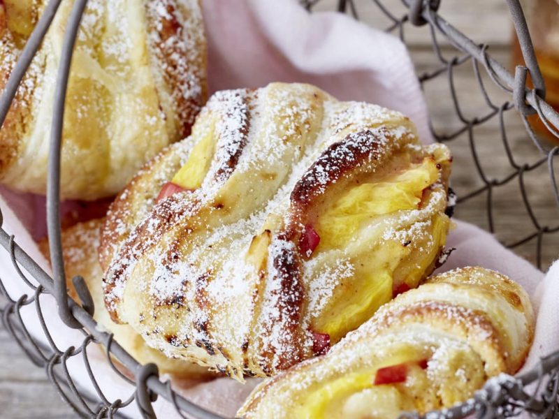 Drei Pfirsich-Schnecken mit Puderzucker auf heller Serviette in einem Metallkörbchen vor Holzhintergrund.