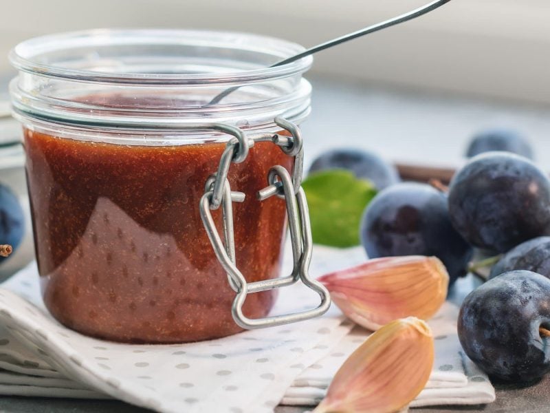 Süß-scharfe Pflaumensauce in Weckglas mit Löffel auf Tuch auf Holz. Daneben Knoblauchzehen und Pflaumen. Frontalansicht.