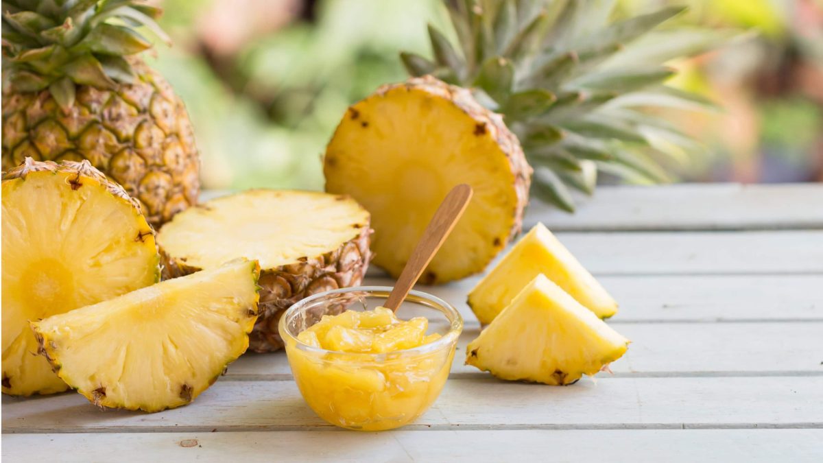 Ein Glas mit der Pina Colada Marmelade auf einem weiß lackierten Holztisch. Daneben einige angeschnittene Ananasfrüchte.