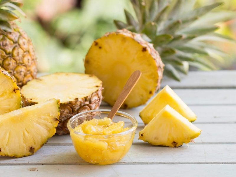 Ein Glas mit der Pina Colada Marmelade auf einem weiß lackierten Holztisch. Daneben einige angeschnittene Ananasfrüchte.