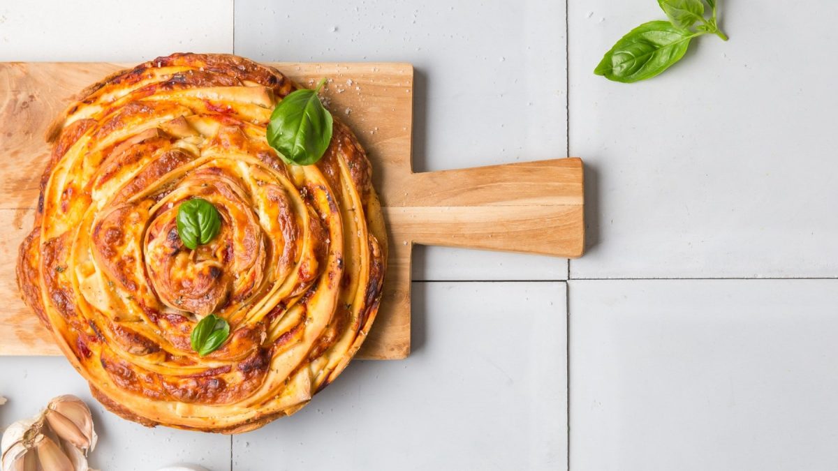 Pizza-Swirl garniert mit Basilikum auf einem Holzbrett vor hellem gefliestem Hintergrund