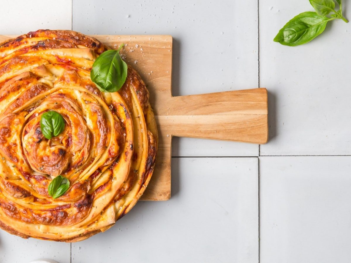 Pizza-Swirl garniert mit Basilikum auf einem Holzbrett vor hellem gefliestem Hintergrund