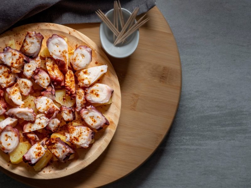 Pulpo a feira mit Kartoffeln auf Holzbrett. Daneben Zahnstocher. Draufsicht.