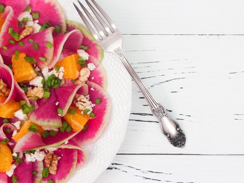 Eine Radieschen-Orangen-Carpaccio auf einem weißen Teller steht auf einem weißen Untergrund aus Holz.