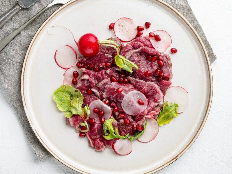 Ein Teller Rinder-Carpaccio mit Radieschen ssteht auf einem hellen Untergrund. Aus der Vogelperspektive.