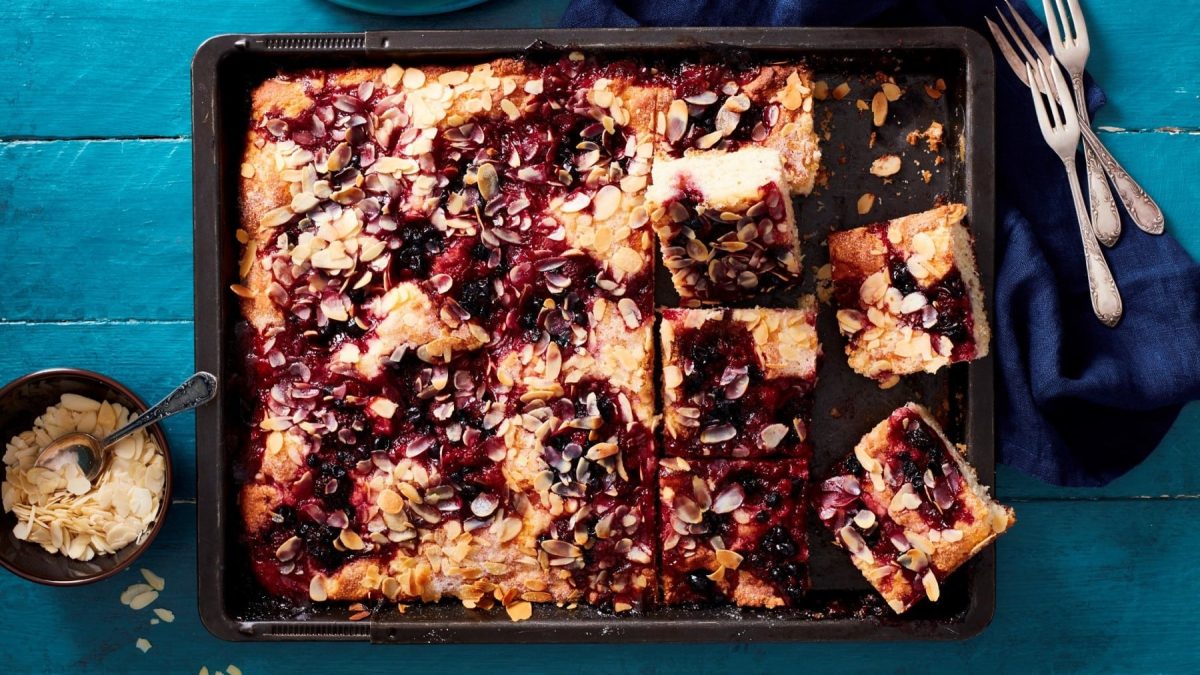 Ein Rote-Grütze-Butterkuchen auf einem Backblech. Daneben liegt Besteck und ein Küchentuch. Außerdem eine Schale mit Mandelblättchen.