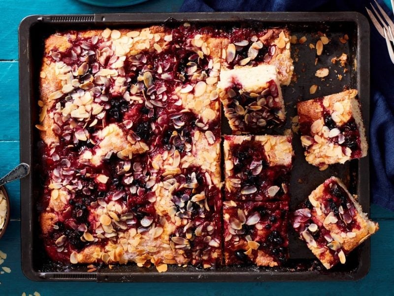 Ein Rote-Grütze-Butterkuchen auf einem Backblech. Daneben liegt Besteck und ein Küchentuch. Außerdem eine Schale mit Mandelblättchen.