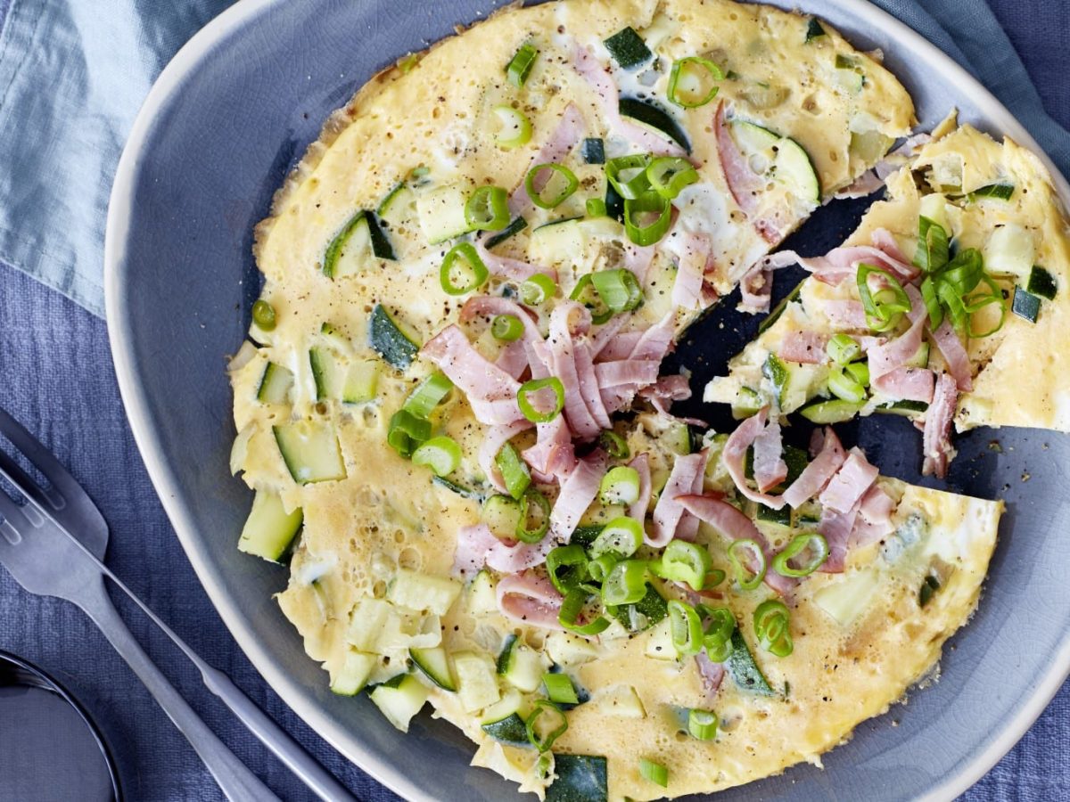 Die Scharfe Tortilla garniert mit Frühlingszwiebeln und Lachsschinken liegt angeschnitten auf einem grauen Teller. Daneben Besteck vor graublauem Hintergrund.