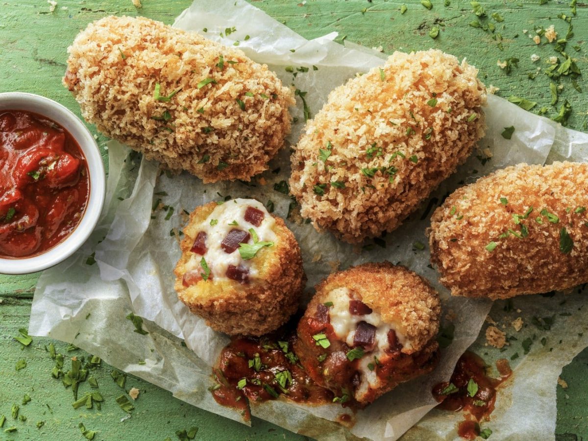 Fünf Schinkenkroketten auf Backpapier garniert mit Petersilie neben einer kleinen Schüssel Tomatensauce vor grünem Hintergrund