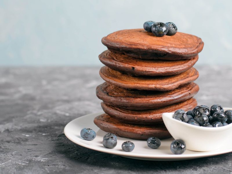 Ein weißer Teller mit den Schokoladen-Pancakes und Blaubeeren auf einem grauen Untergrund.