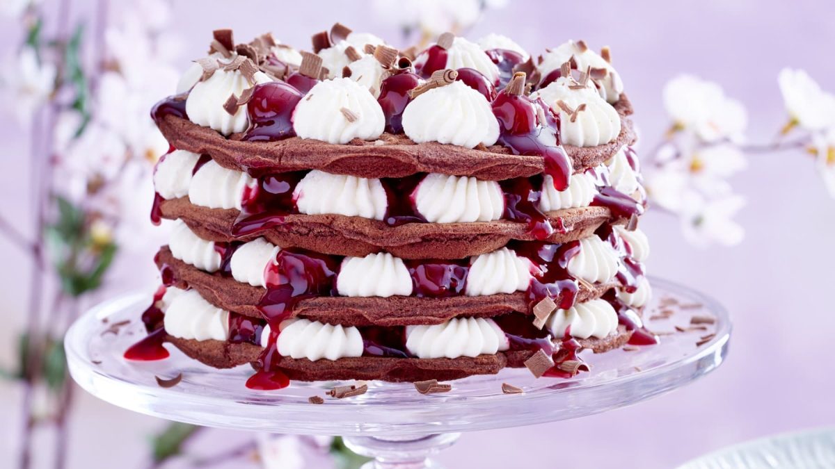 Schwarzwäler-Kirsch-Waffeltorte garniert mit Schokoraspeln auf einem Glas-Tortenteller vor Blütenhintergrund.