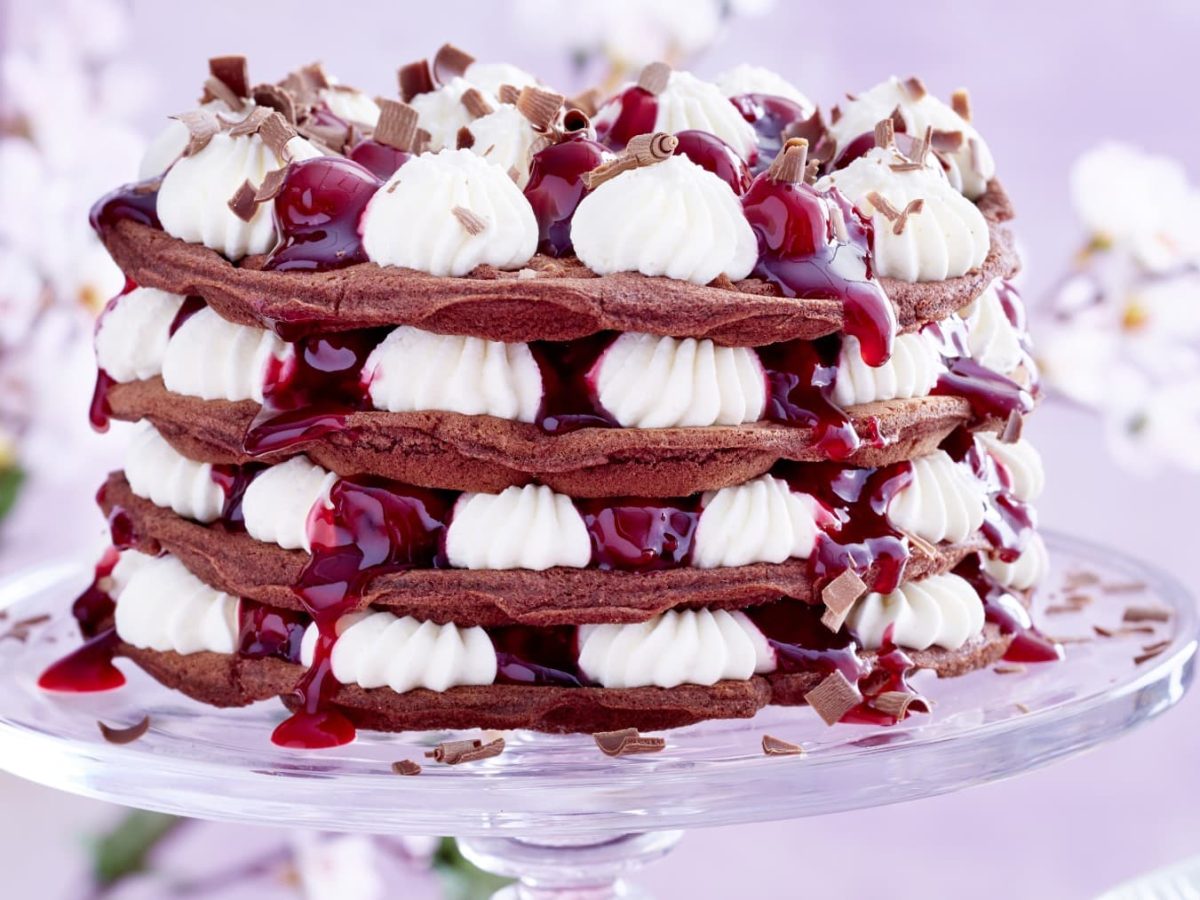 Schwarzwäler-Kirsch-Waffeltorte garniert mit Schokoraspeln auf einem Glas-Tortenteller vor Blütenhintergrund.