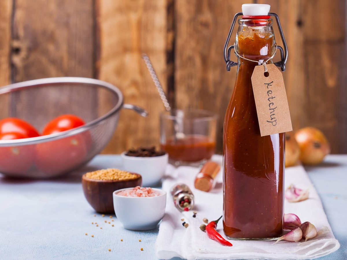 Selbst gemachter Curry Ketchup in Flasche mit Etikett auf weißem Tuch und blauem Untergrund. Im Hintergrund Gewürze, Tomaten in einem Sieb, Zwiebeln, Chili, Knoblauch und Glas mit Sauce und Löffel. Frontalansicht.