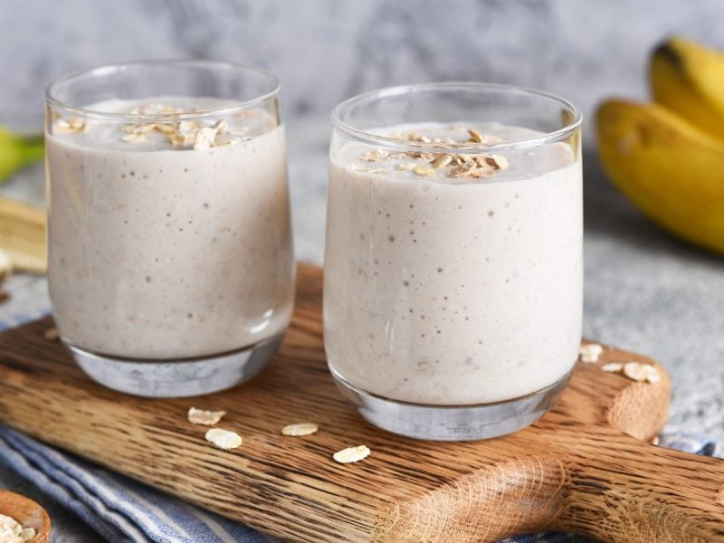 Zwei Gläser mit Bananen-Hafer-Smoothie auf einem Holzbrett neben frischen Bananen