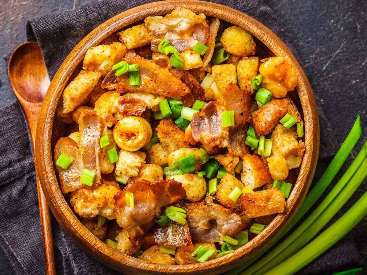 Ein Holzteller mit Migas de Pastor auf einem dunklen Untergrund und einer Stange Lauch.