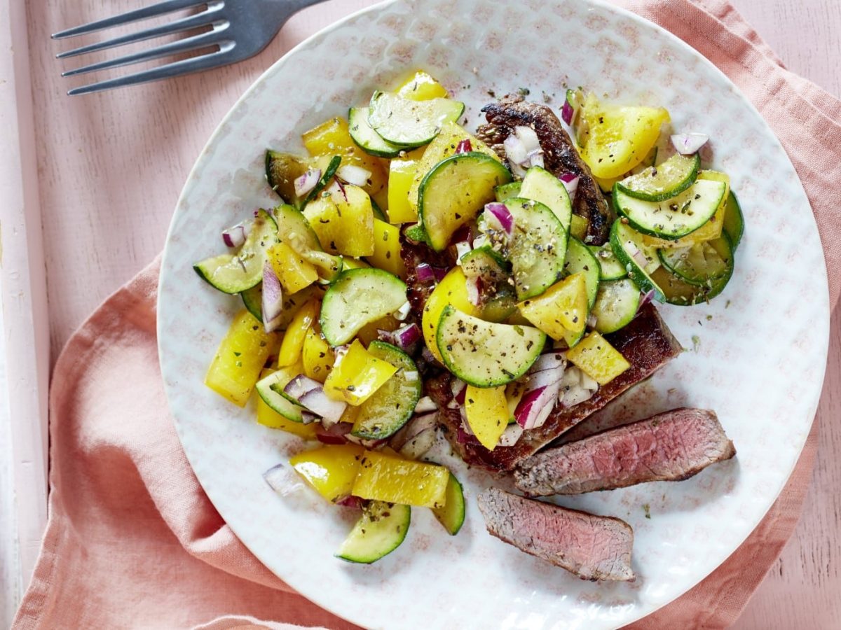 In Streifen geschnittenes Steak mit Mittelmeergemüse also Paprika und Zucchini auf einem runden Teller vor hellem Hintergrund