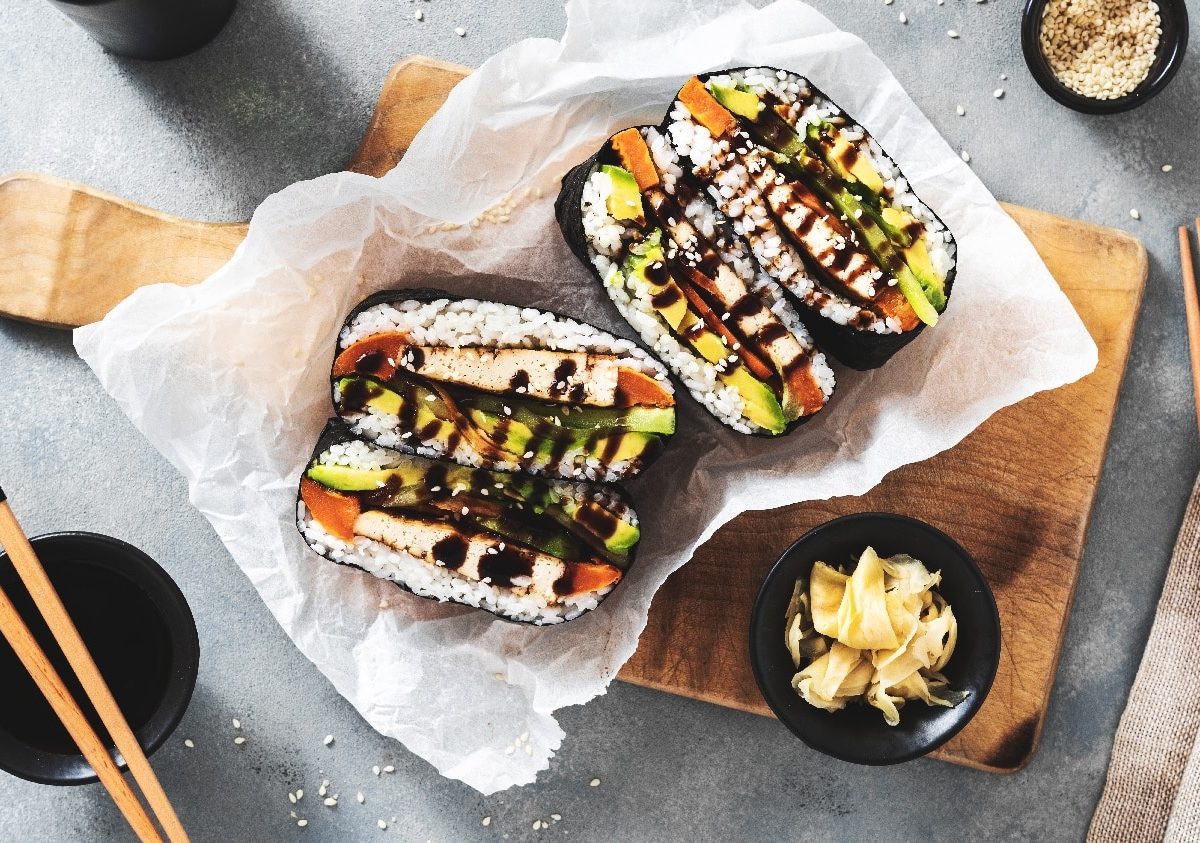 Ein aufgeschnittenes Sushi-Sandwich auf einem kleinen Holzbrett mit Essstäbchen, Sojasauce und Sesam.