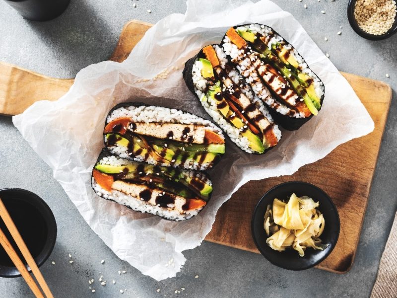 Ein aufgeschnittenes Sushi-Sandwich auf einem kleinen Holzbrett mit Essstäbchen, Sojasauce und Sesam.