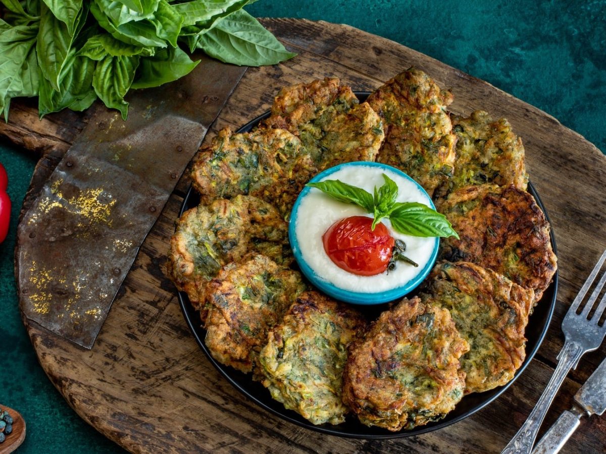 Ein Teller Brokkoli-Kartoffelpuffer, in deren Mitte eine Schale mit weißem Dip steht, daneben Besteck und frisches Gemüse.