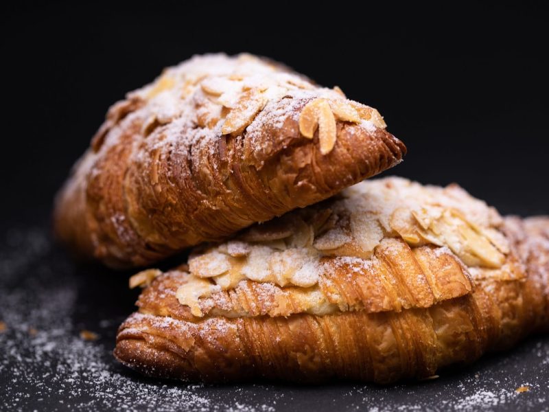 zwei vegane Mandel-Croissants mit Puderzucker in dunklem Setting.