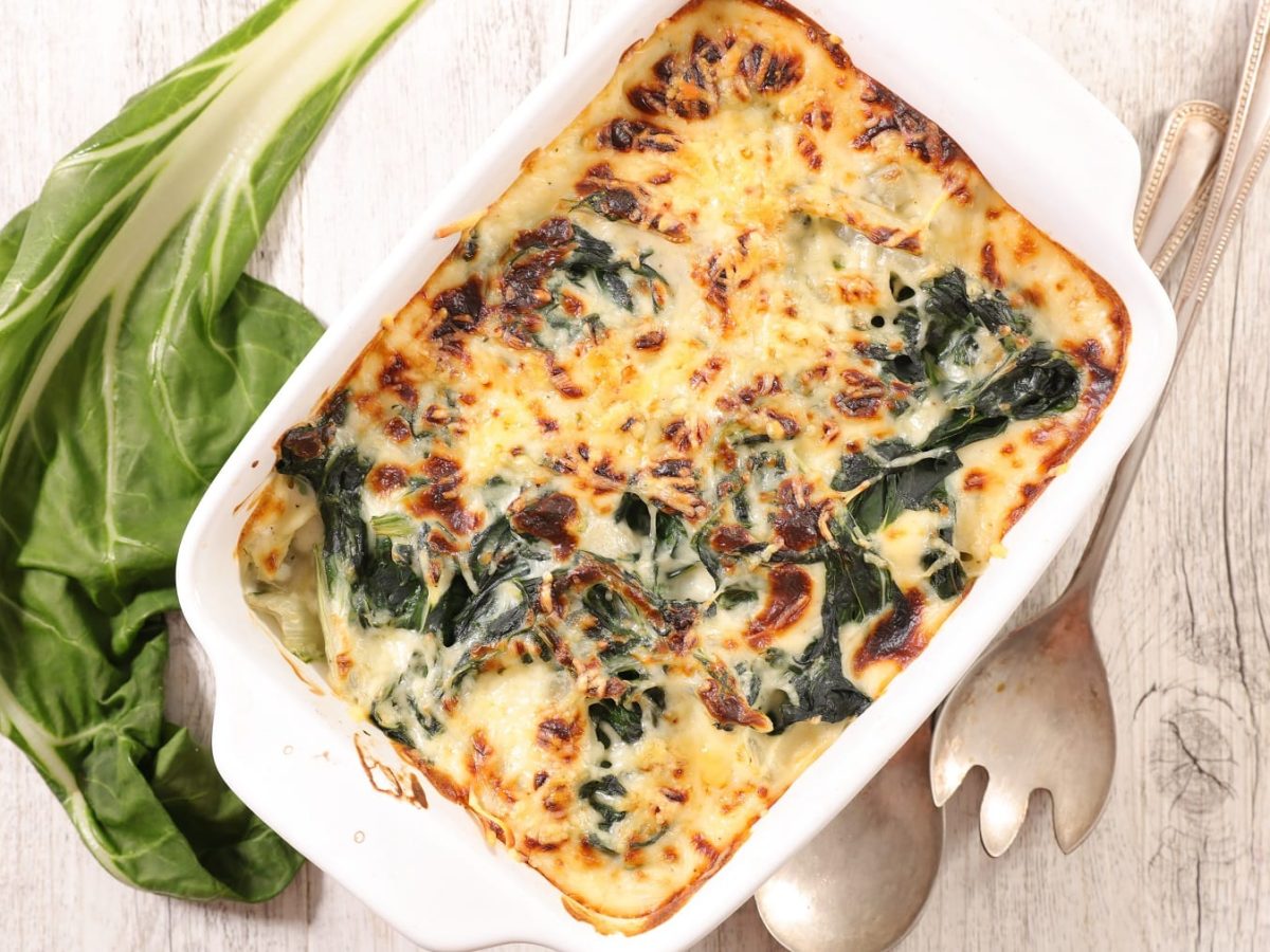 Eine vegane Mangold-Lasagane in der Auflaufform in der Draufsicht, daneben frischer Mangold und Besteck.