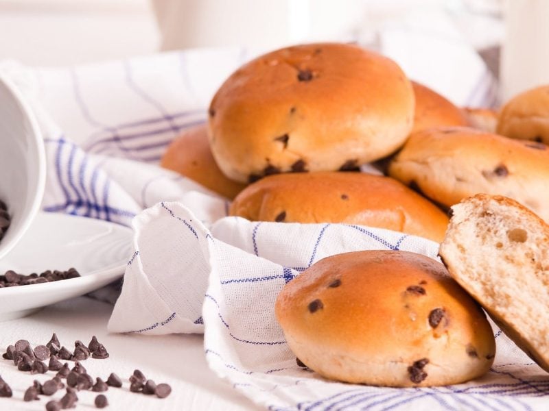 Vegane Schokobrötchen, eins davon angeschnitten, auf einem karierten Geschirrtuch, daneben eine umgekippte Schüssel mit Schokotropfen.