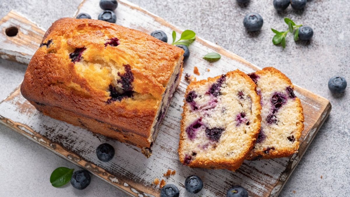 Ein angeschnittener Blaubeer-Zitronenkuchen (Kastenform) auf einem Brett, daneben liegen frische Blaubeeren.