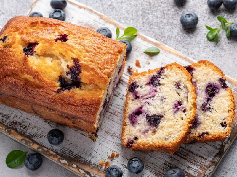 Ein angeschnittener Blaubeer-Zitronenkuchen (Kastenform) auf einem Brett, daneben liegen frische Blaubeeren.