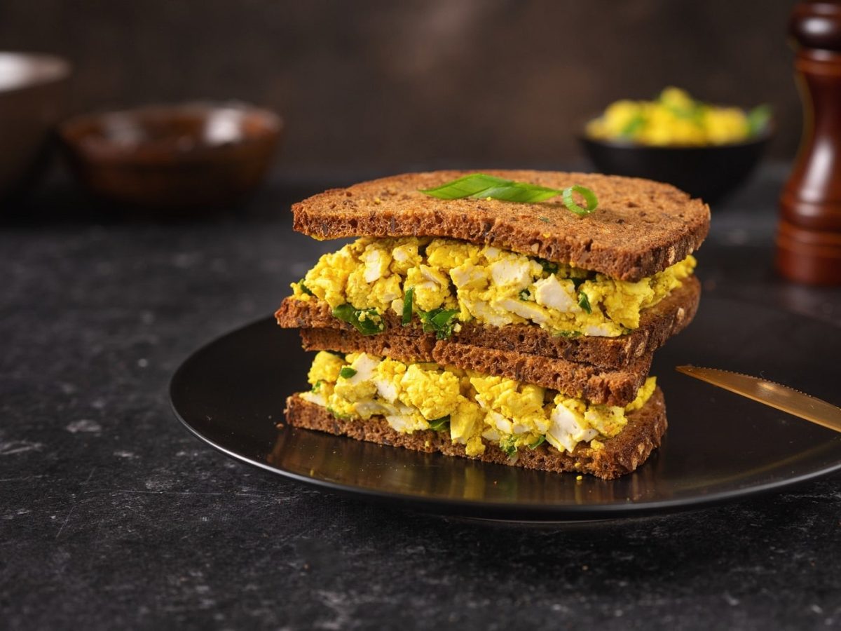 Zwei Sandwiches mit dunklem Brot und veganem Eiersalat gestapelt auf einem Teller, daneben liegt ein goldenes Messer.