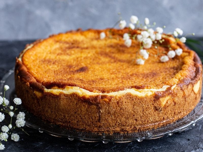 Ein veganer Mango-Vanille-Cheesecake auf einem Tortenteller aus Glas, mit weißen Blüten verziert.