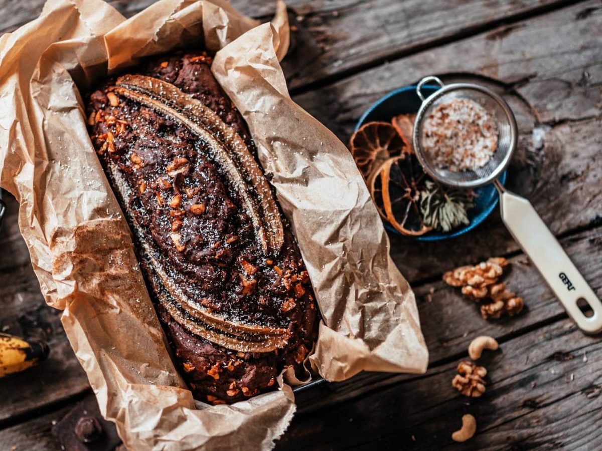 Ein veganes Schoko-Bananenbrot mit Walnüssen und einer halbierten Banane in deiner Kastenform mit Backpapier, daneben Schalen mit Zutaten, alles Draufsicht.