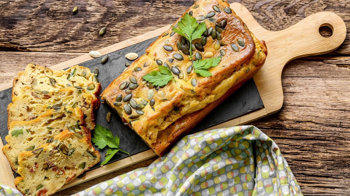 Ein angeschnittenes veganes süßes Zucchinibrot mit Kürbiskernen auf einem Holzbrett in der Draufsicht.