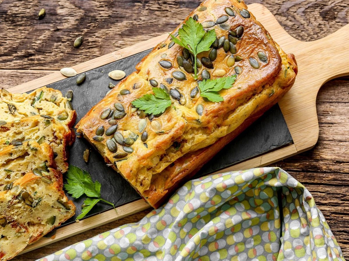 Ein angeschnittenes veganes süßes Zucchinibrot mit Kürbiskernen auf einem Holzbrett in der Draufsicht.