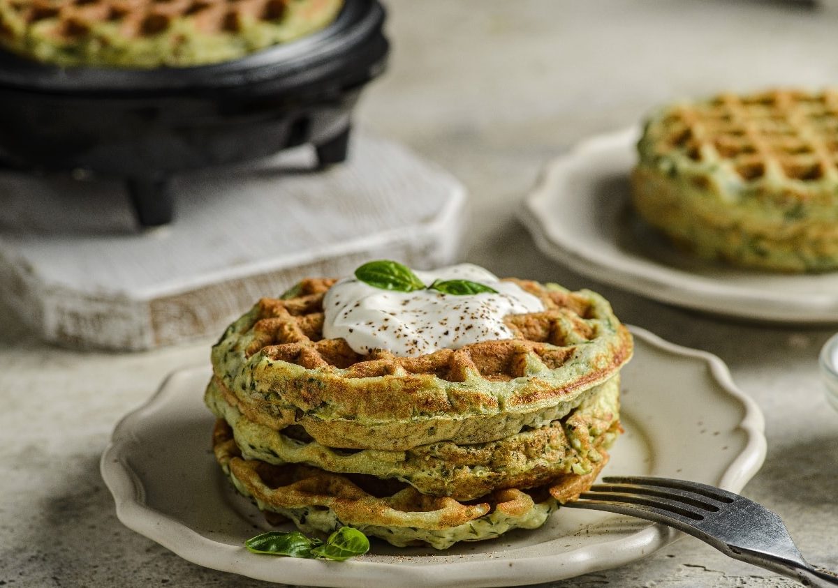 Zwei beige Teller mit jeweils zwei Zucchini-Waffeln auf einem beigen Tisch. Auf dem vorderen Teller ist außerdem ein Stück Burrata in der Mitter der Waffeln platziert.