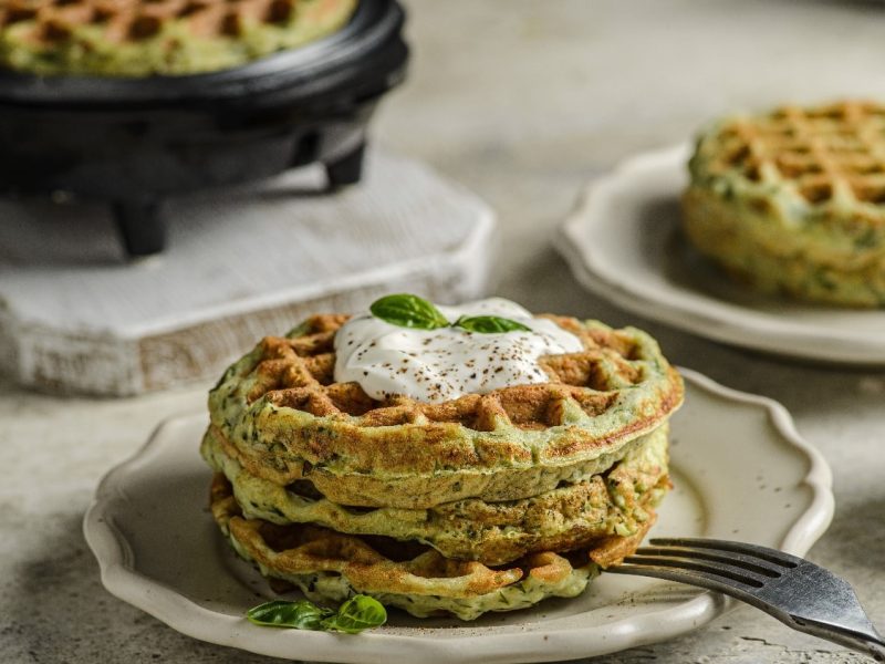 Zwei beige Teller mit jeweils zwei Zucchini-Waffeln auf einem beigen Tisch. Auf dem vorderen Teller ist außerdem ein Stück Burrata in der Mitter der Waffeln platziert.