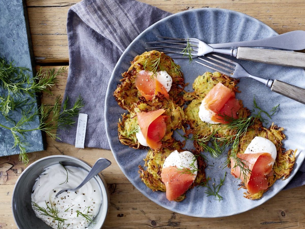 Zucchinipuffer mit Räucherlachs auf einem hellblauen Teller neben Dill und Schale mit saurer Sahne vor Holzhintergrund.