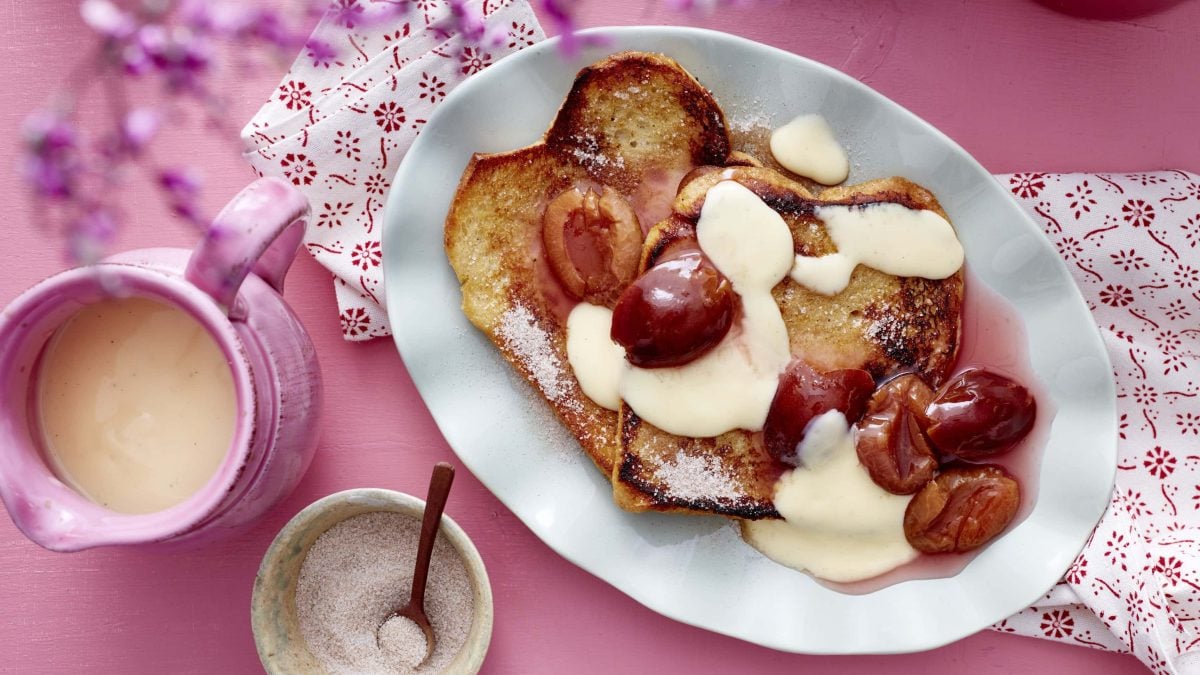 Arme Ritter mit Pflaumenkompott und Vanillesauce, serviert auf einem länglichen Teller auf rosanem Untergrund.