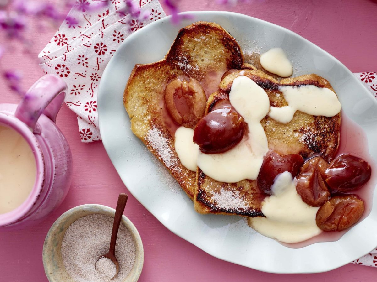 Arme Ritter mit Pflaumenkompott und Vanillesauce, serviert auf einem länglichen Teller auf rosanem Untergrund.