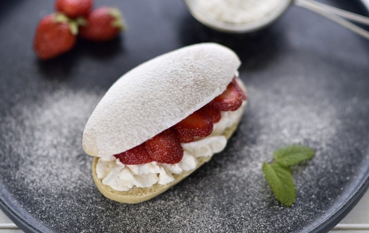 Auf einem Teller liegt ein veganer Brioche-Bun. Er is tmit Sahne und Erdbeeren gefüllt und mit Puderzucker bestreut.