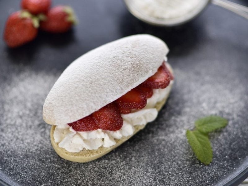 Auf einem Teller liegt ein veganer Brioche-Bun. Er is tmit Sahne und Erdbeeren gefüllt und mit Puderzucker bestreut.