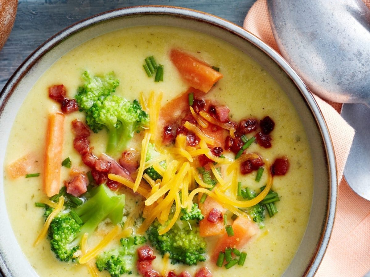 Cremige Cheddar-Brokkoli-Suppe, garniert mit Speck, frischem Brokkoli und Schnittlauch, serviert in einer Tonschale auf einem grauen Holztisch.