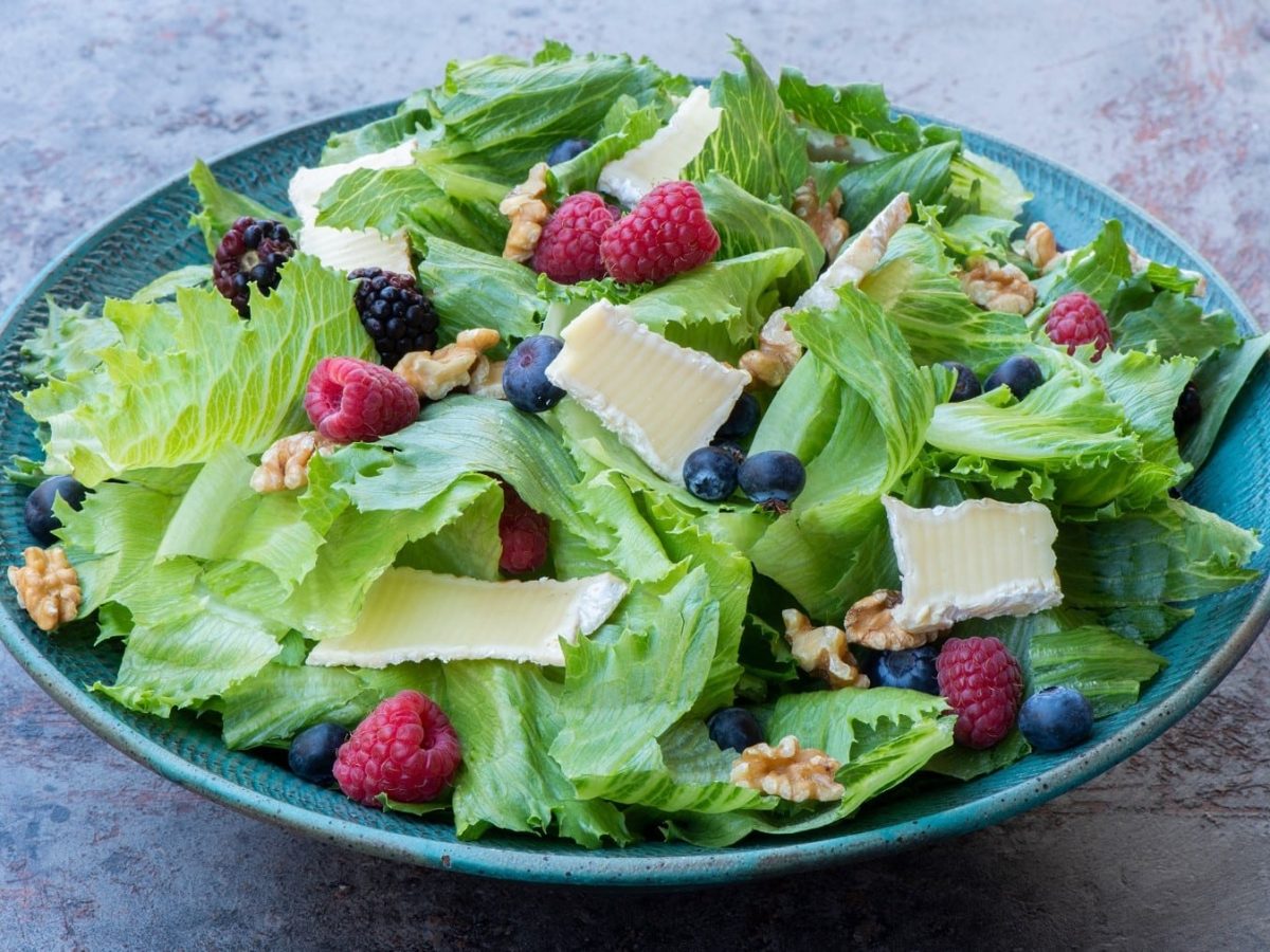 Endivien-Salat mit Beeren und Brie, garniert mit Walnüssen und saurer Vinaigrette und serviert in einer großen, blauen Tonschale auf einem hellgrauen Schiefertisch