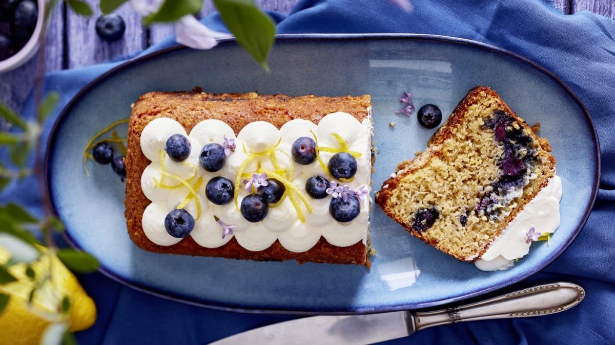 Auf einem Teller liegt ein angeschnittener Heidelbeer-Haferflocken-Kuchen. Er ist mit Zitronenzesten und Blüten verziert.