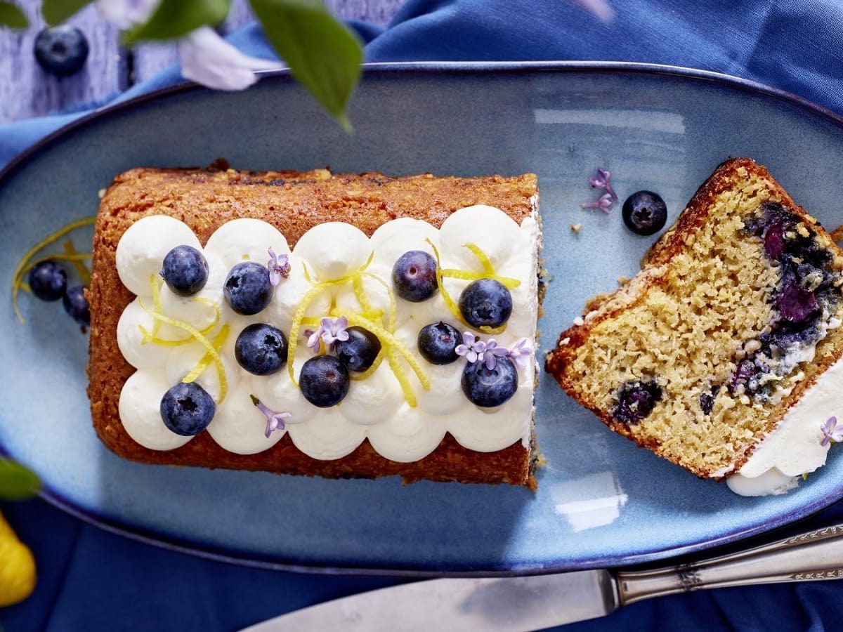 Auf einem Teller liegt ein angeschnittener Heidelbeer-Haferflocken-Kuchen. Er ist mit Zitronenzesten und Blüten verziert.